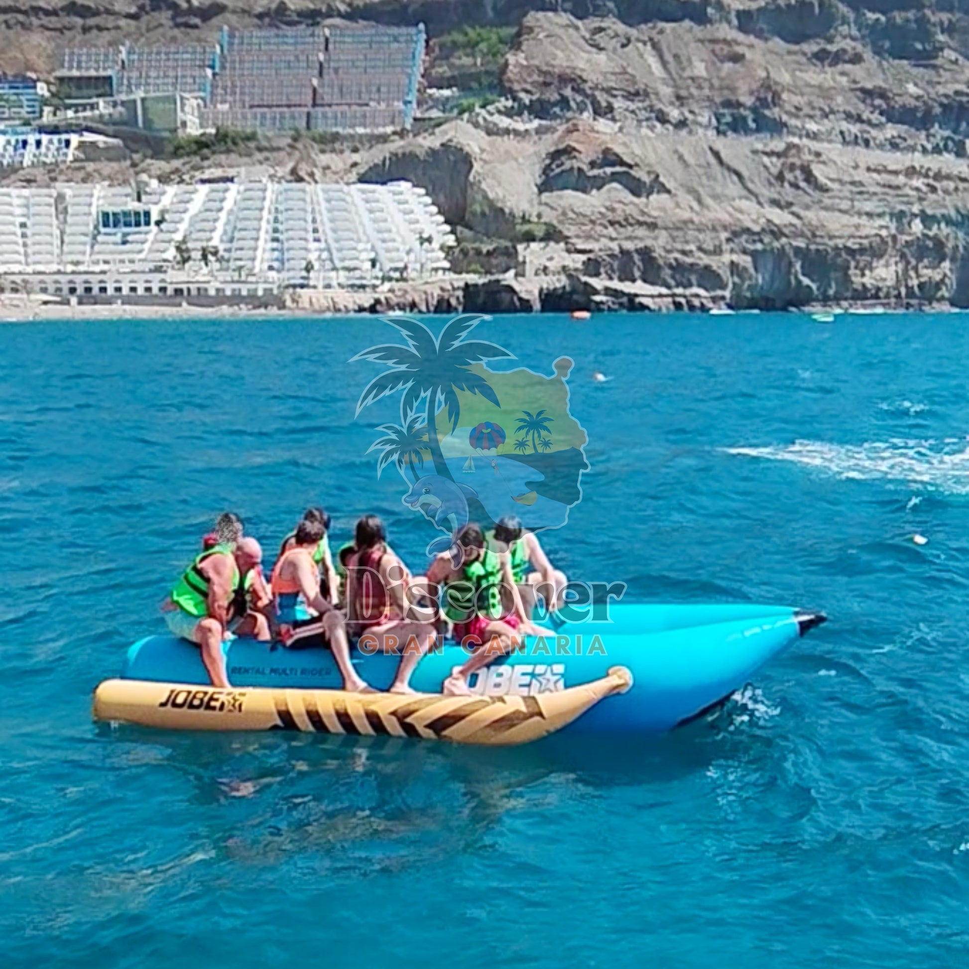 Group of people on a banana boat in clear blue water with cliffs and buildings in the background.