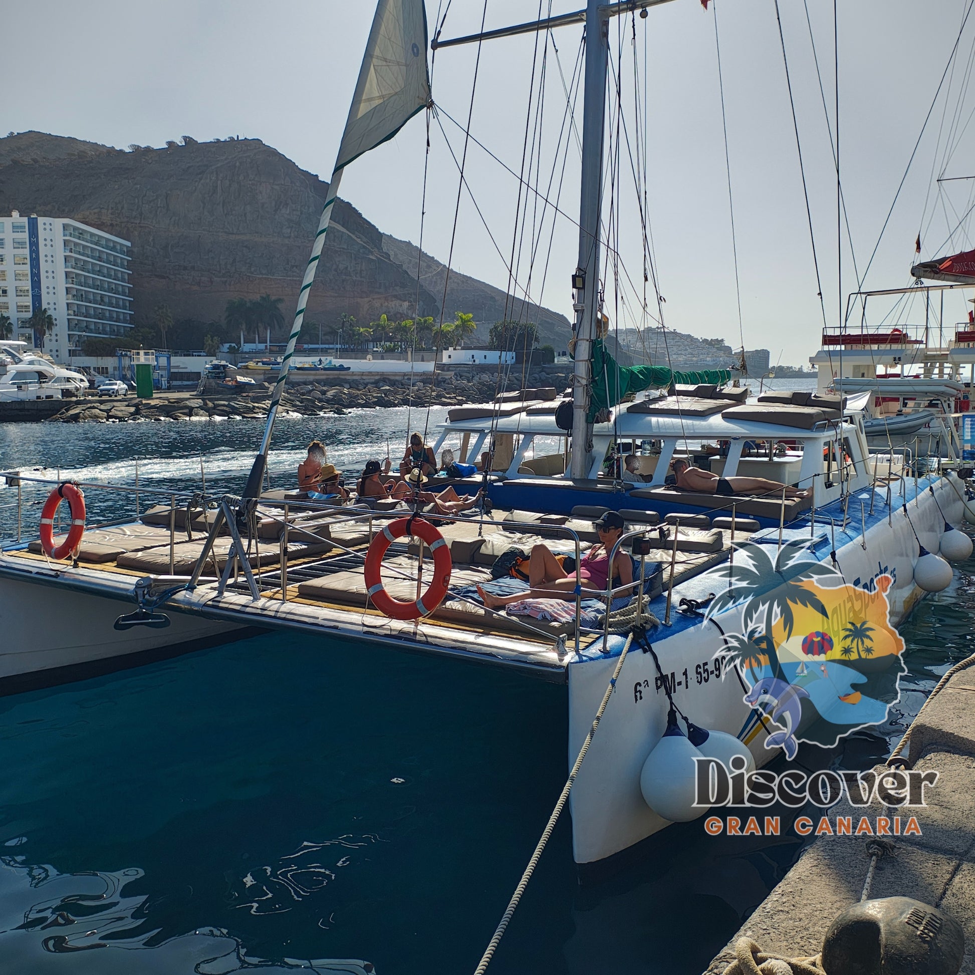 Catamaran magic 3 docked at a harbor with people on board, mountains in the background.