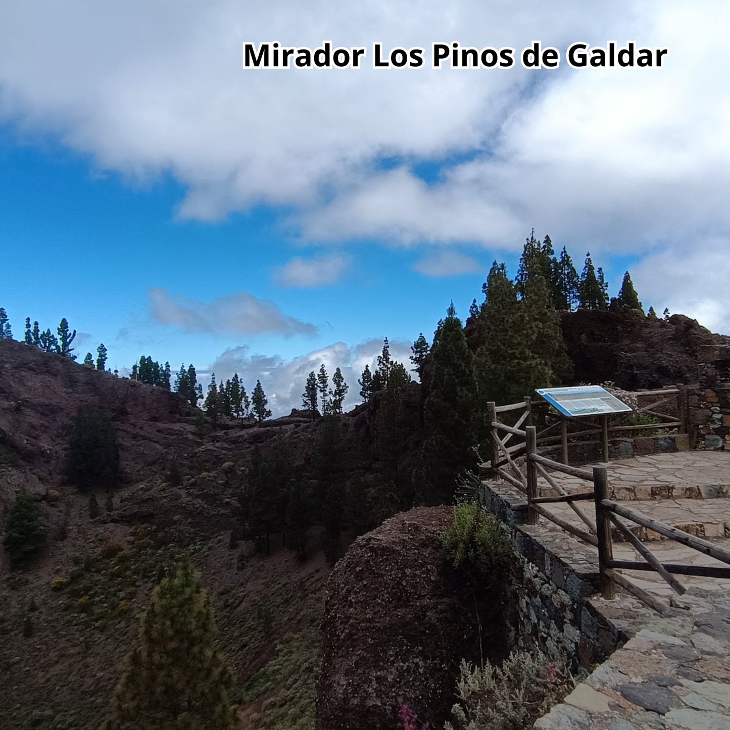 Scenic view of Mirador Los Pinos de Galdar with a wooden platform and sign, surrounded by trees and mountains.