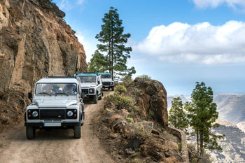 Jeep safari convoy driving along a narrow mountain road in Gran Canaria with dramatic cliffs, pine trees and panoramic Atlantic Ocean views.