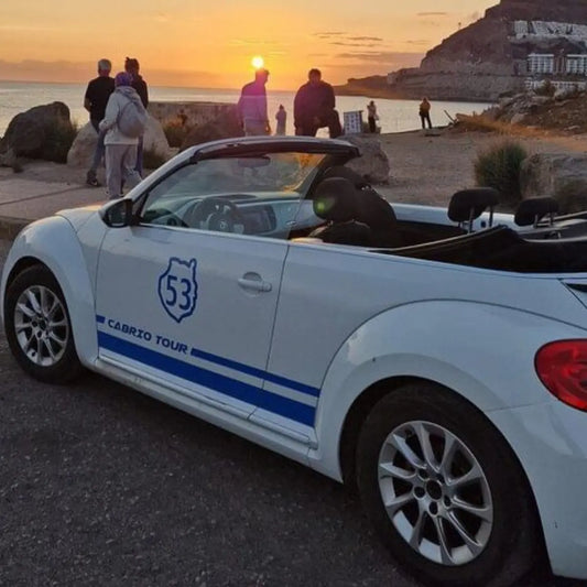 White convertible car with blue stripes and '53 Cabrio Tour' text parked on a scenic road at sunset.