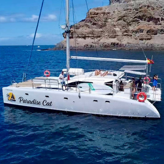 White catamaran named 'Paradise Cat' on the water with people around, cliffs in the background.