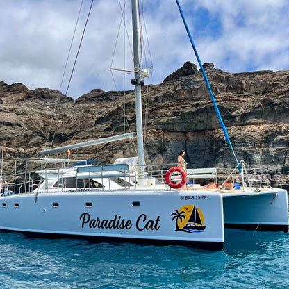 White catamaran named 'Paradise Cat' on the water with a rocky cliff in the background