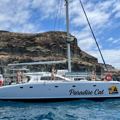 White catamaran named 'Paradise Cat' with people on board, set against a rocky cliff and blue sky.