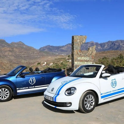 Two convertible cars, one blue and one white with blue stripes, parked in front of a mountainous landscape of Mirador La Sorrueda in Gran Canaria