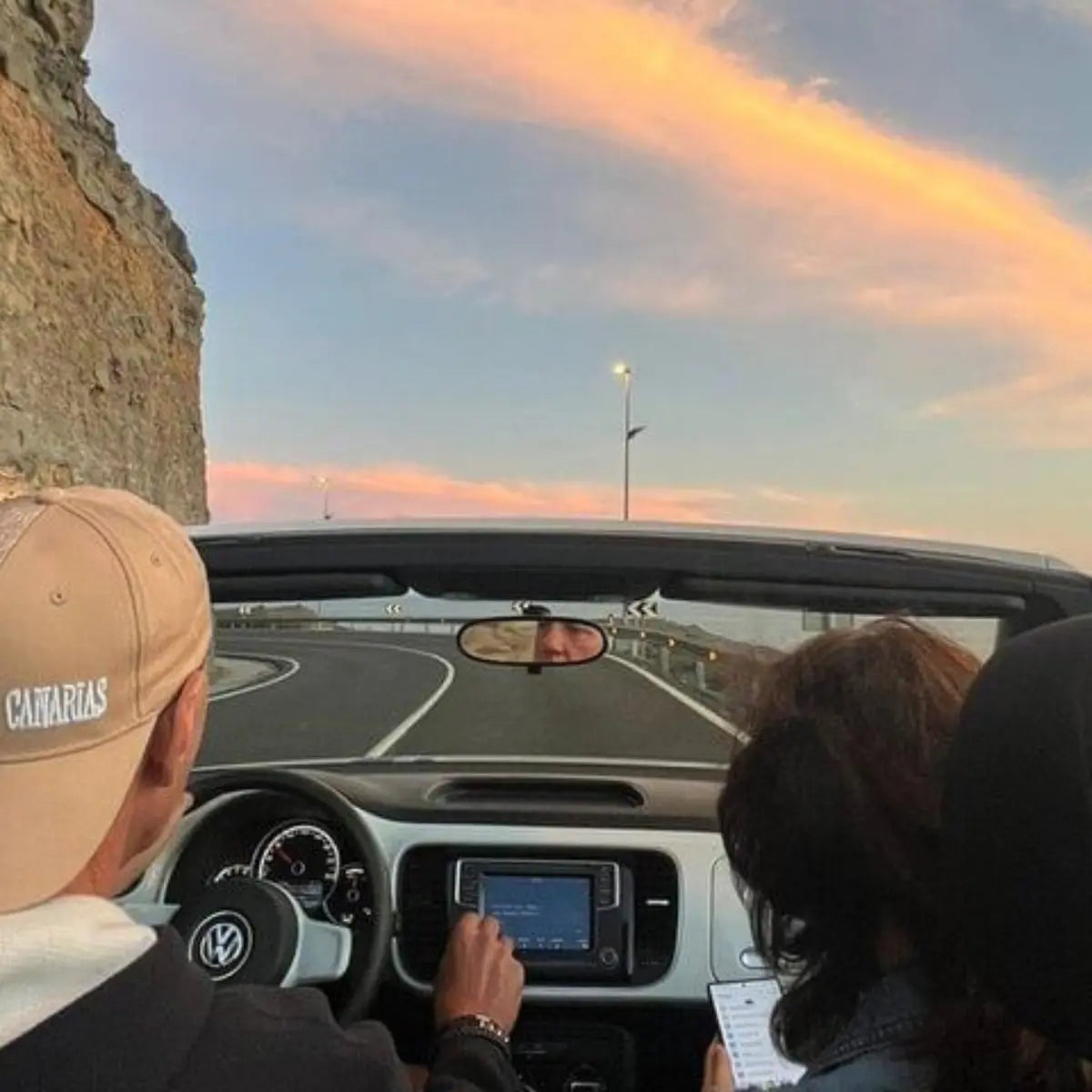 Two people in a convertible car with a scenic road and sunset sky.