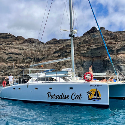 White catamaran named 'Paradise Cat' with people on board, surrounded by rocky cliffs and blue sky.