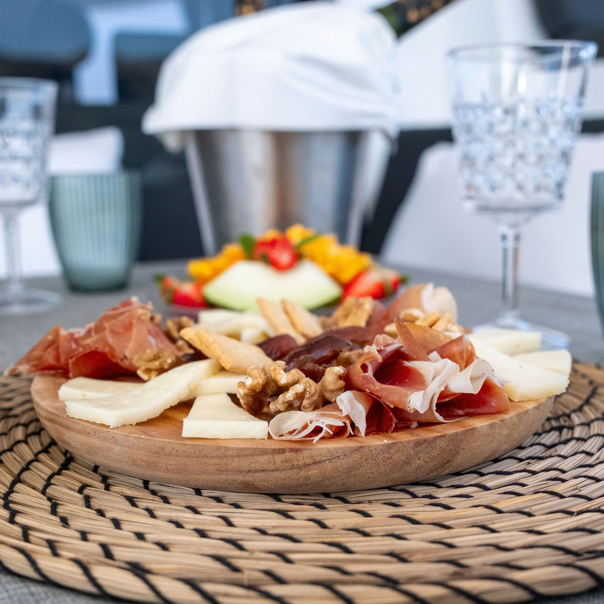 A wooden charcuterie board filled with sliced cured meats, cheese, crackers, and nuts, set on a woven placemat at a dining table with blurred glasses and a bucket in the background.