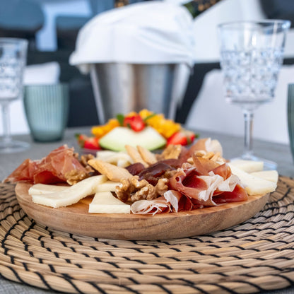 A wooden charcuterie board filled with sliced cured meats, cheese, crackers, and nuts, set on a woven placemat at a dining table with blurred glasses and a bucket in the background.