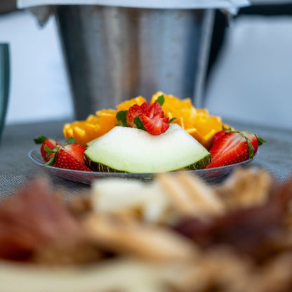A close-up of a fruit platter with a slice of cantaloupe melon, strawberries, and yellow fruit pieces in the foreground, with a blurred metal bucket in the background.