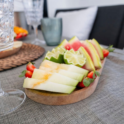 A wooden serving board filled with sliced melon, lime wedges, strawberries, and watermelon on a dining table with glassware in the background.