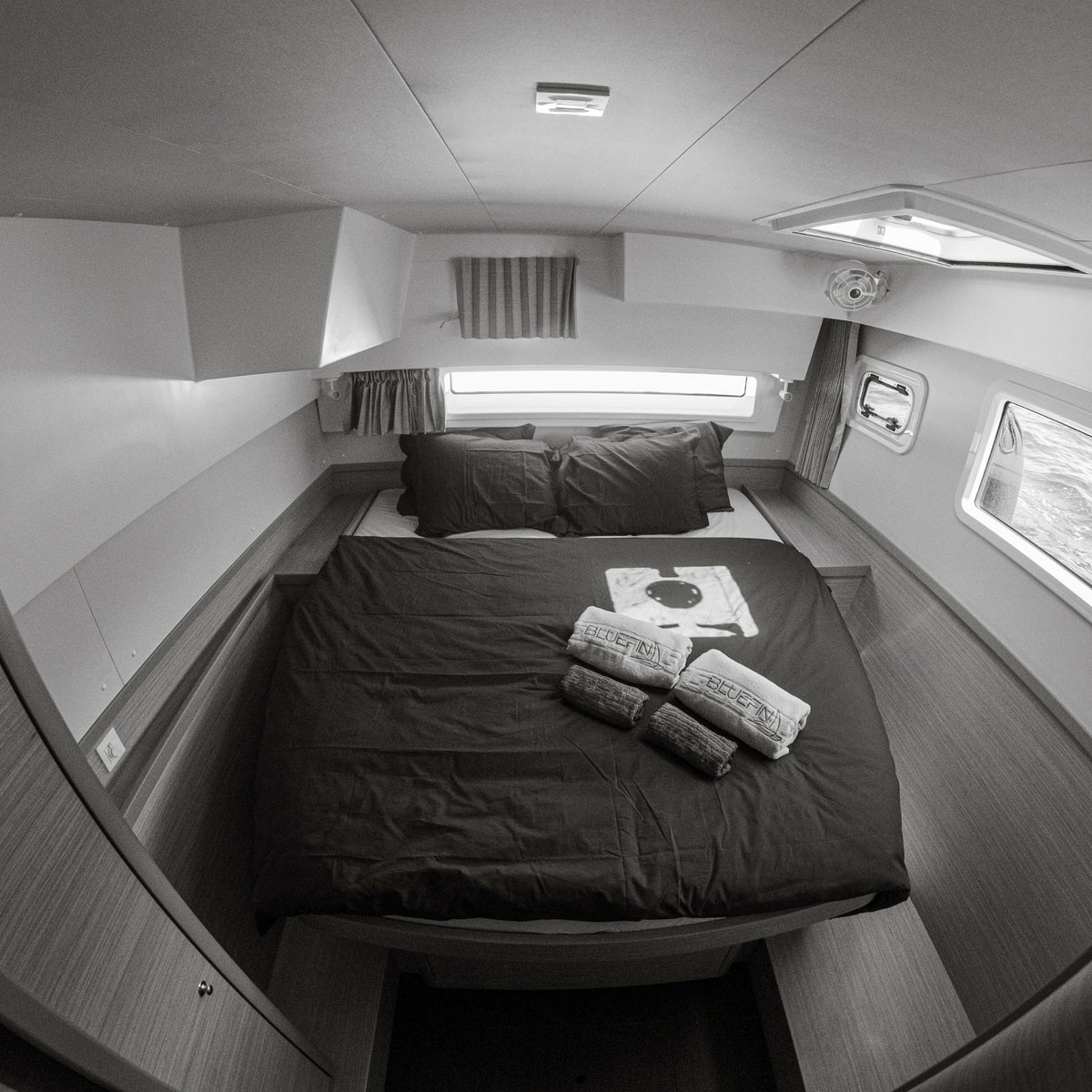 Bluefin catamaran: interior view of a compact bed in a boat or cabin, with dark bedding, pillows, curtains, windows, and overhead lighting visible.