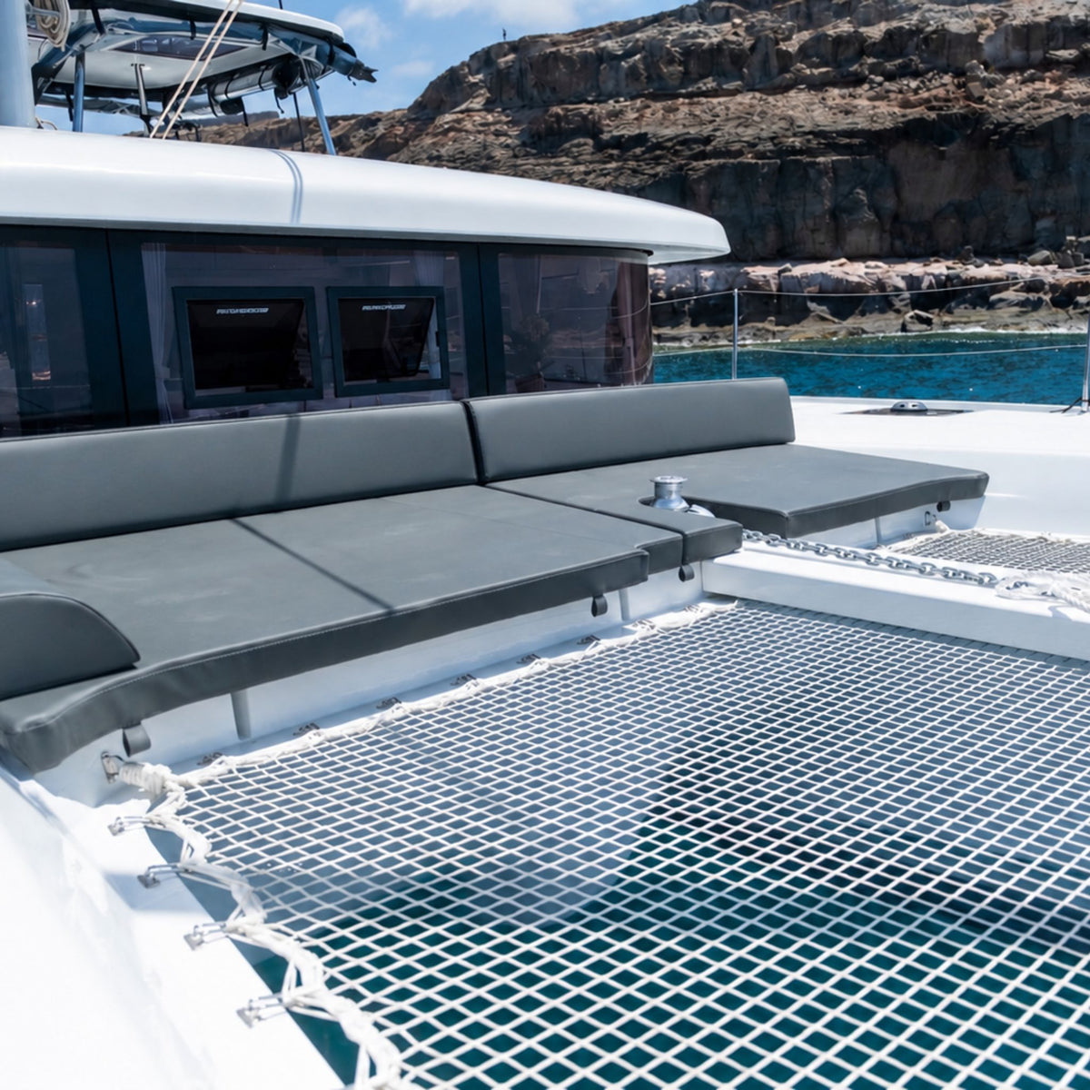 A close-up view of a bluefin catamaran gran canaria deck featuring a gray mesh floor, cushioned gray seating along the side, and an outdoor setting with rocky cliffs and blue water in the background.
