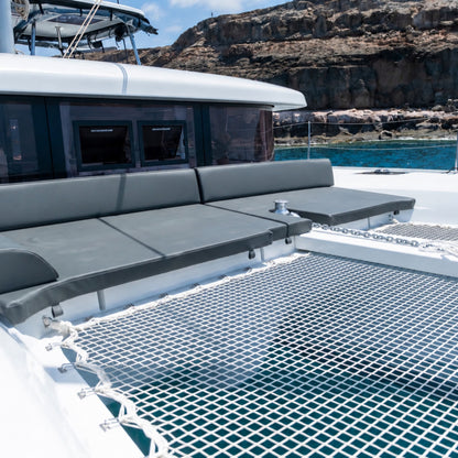 A close-up view of a bluefin catamaran gran canaria deck featuring a gray mesh floor, cushioned gray seating along the side, and an outdoor setting with rocky cliffs and blue water in the background.