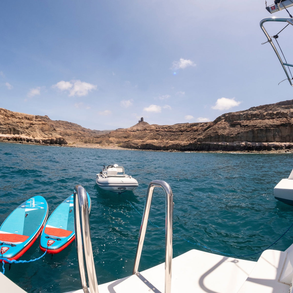 A scenic view from a boat showing turquoise blue water, rocky brown cliffs, and a small white motorboat in the distance under a clear blue sky. Two bright blue and red paddleboards are docked in the foreground on the left, with the white boat railing and ladder visible at the bottom.