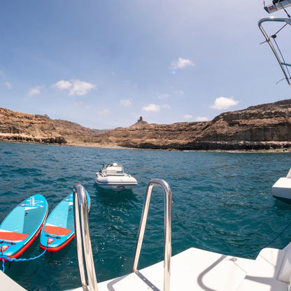 A scenic view from a boat showing turquoise blue water, rocky brown cliffs, and a small white motorboat in the distance under a clear blue sky. Two bright blue and red paddleboards are docked in the foreground on the left, with the white boat railing and ladder visible at the bottom.