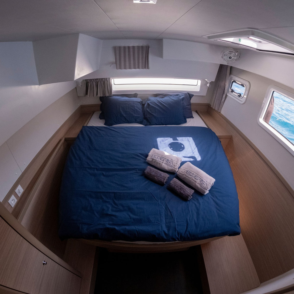 Bluefin catamaran puerto de mogan: compact double bed with navy blue bedding and pillows inside a boat cabin, with three rolled towels and a small towel bar placed on top. The room has wood-look paneling, white walls, and windows showing water outside.