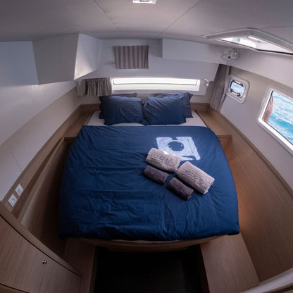Bluefin catamaran puerto de mogan: compact double bed with navy blue bedding and pillows inside a boat cabin, with three rolled towels and a small towel bar placed on top. The room has wood-look paneling, white walls, and windows showing water outside.