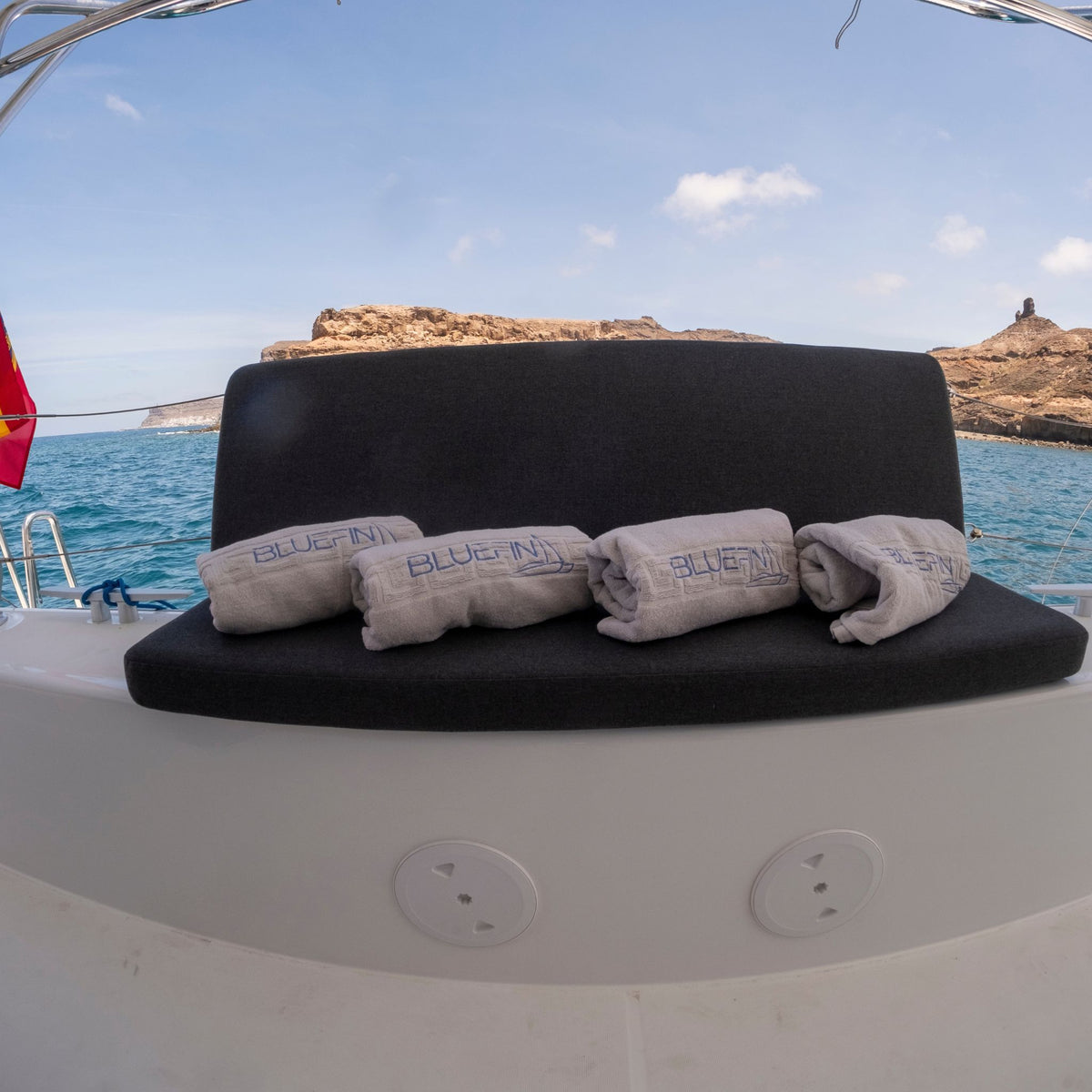 A black upholstered boat bench seat with four rolled light gray towels printed with the brand name 'BLUE FIN PUERTO DE MOGAN' resting on it, set against an outdoor ocean backdrop with rocky islands in the distance.