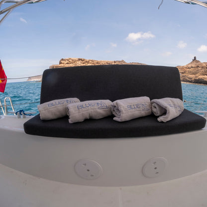 A black upholstered boat bench seat with four rolled light gray towels printed with the brand name 'BLUE FIN PUERTO DE MOGAN' resting on it, set against an outdoor ocean backdrop with rocky islands in the distance.