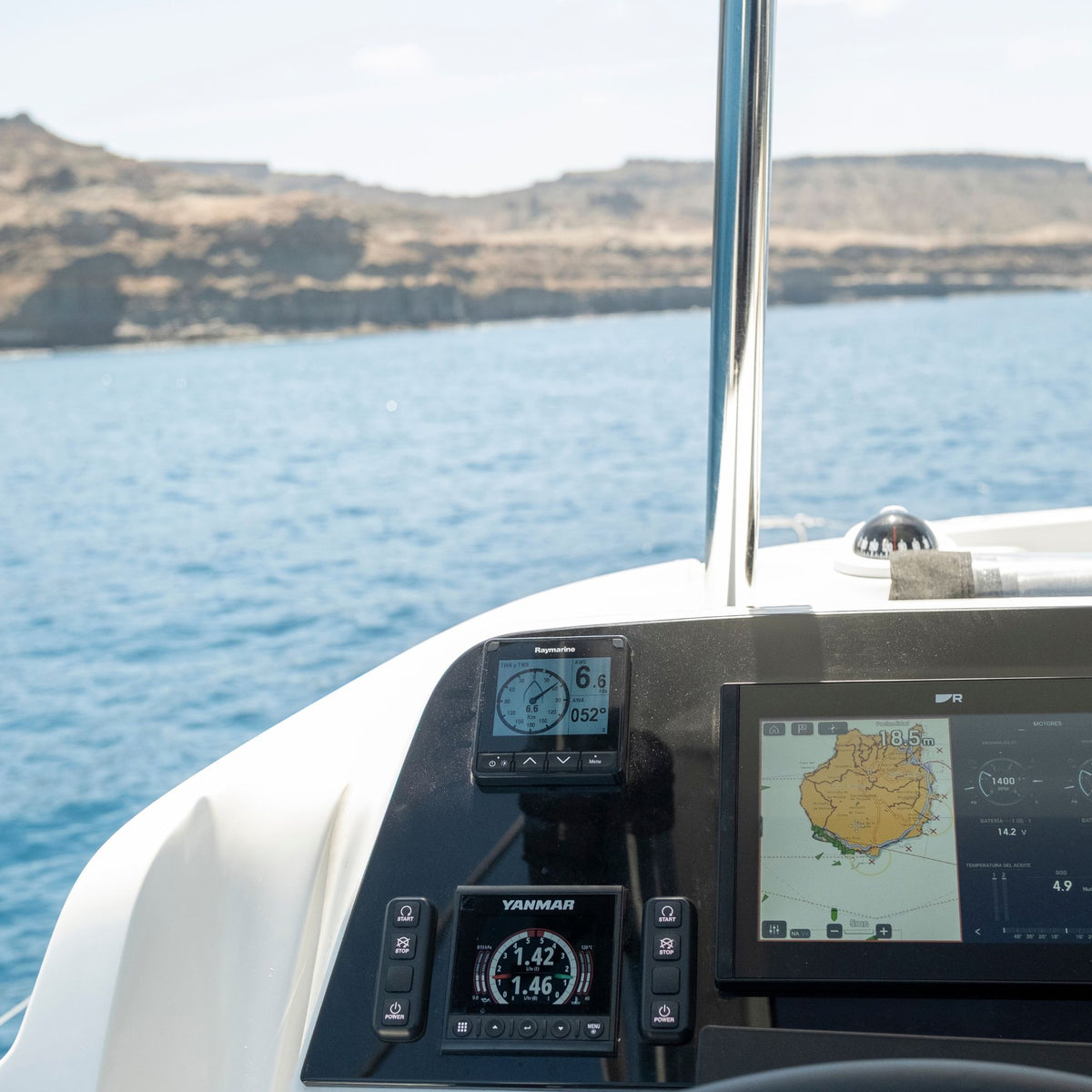 A close-up of a boat's navigation console featuring a large touchscreen display and two smaller Garmin GPS devices mounted on a black panel, with blue water and rocky cliffs in the background and the map of Gran Canaria.