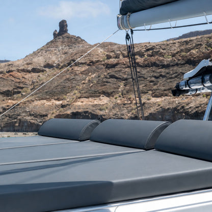 Bluefin catamaran puerto de mogan: close-up of a dark gray boat sun pad or cover with padded sections, attached to a white boat structure, with a rocky brown mountain landscape and blue sky in the background.