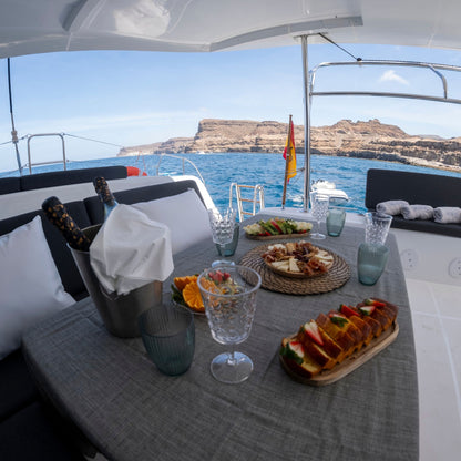 A luxury yacht dining table set with a bottle of champagne in an ice bucket, glasses, sliced bread, and fruit, with a scenic blue sea and rocky coastline in the background.