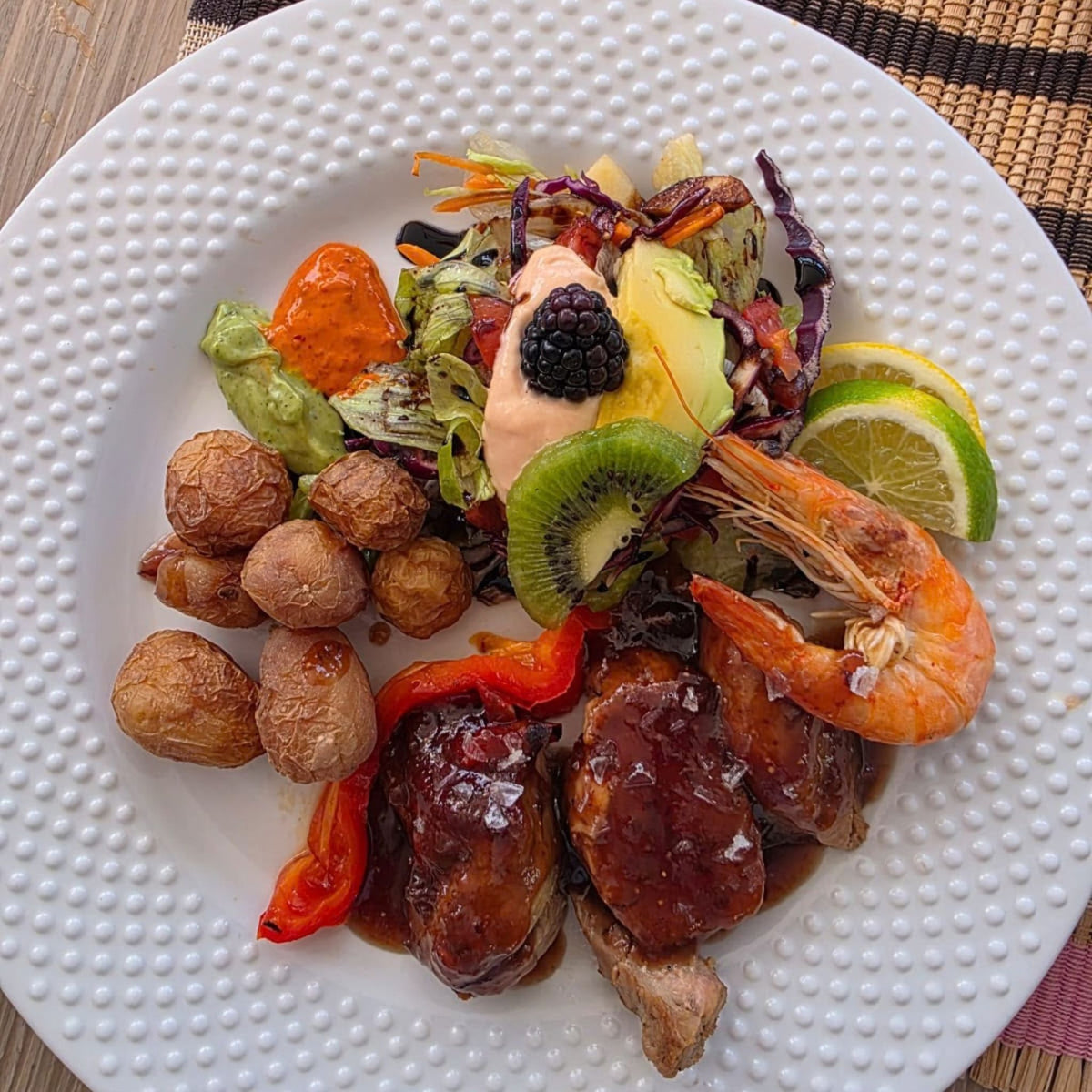 Lunch on the Bluefin catamaran Gran Canaria: a plated prepared food dish featuring glazed shrimp, small roasted potatoes, red peppers, a mixed salad with avocado, kiwi, lime, and a blackberry on top, served on a white textured plate.