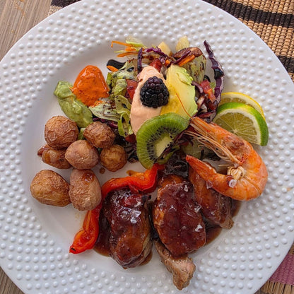 Lunch on the Bluefin catamaran Gran Canaria: a plated prepared food dish featuring glazed shrimp, small roasted potatoes, red peppers, a mixed salad with avocado, kiwi, lime, and a blackberry on top, served on a white textured plate.