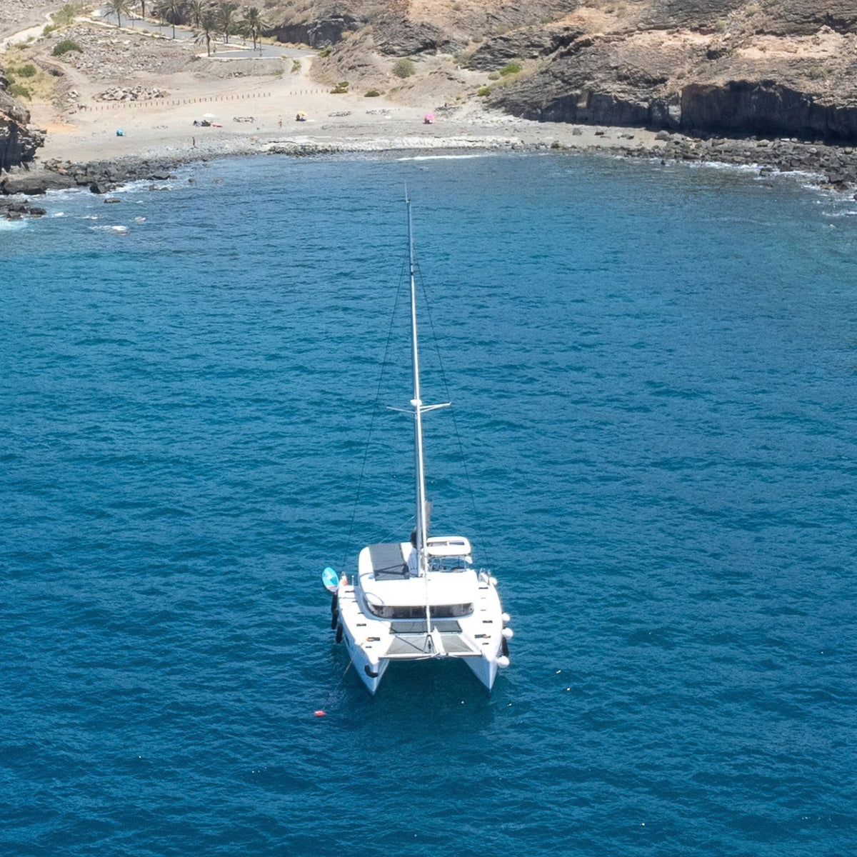 The bluefin catamaran with its mast raised is anchored in deep blue ocean water of Gran Canaria, near a rocky coastal bay (veneguera beach) with a sandy beach and cliffs in the background.