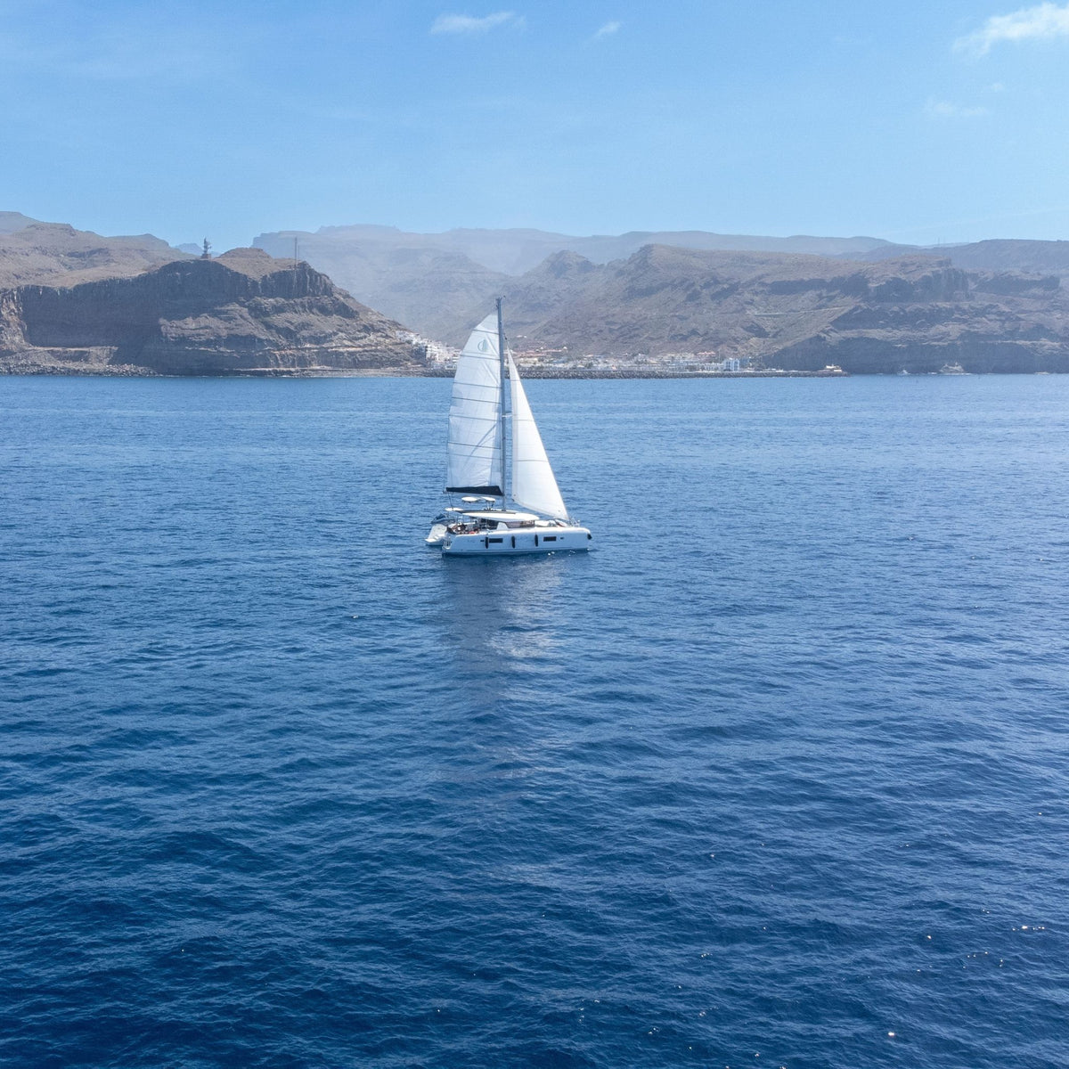 The bluefin catamarann with tall white sails is centered on deep blue ocean water of Gran Canaria's west coast, with rocky mountains and a pale blue sky in the background.