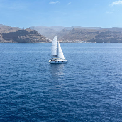 The bluefin catamarann with tall white sails is centered on deep blue ocean water of Gran Canaria's west coast, with rocky mountains and a pale blue sky in the background.